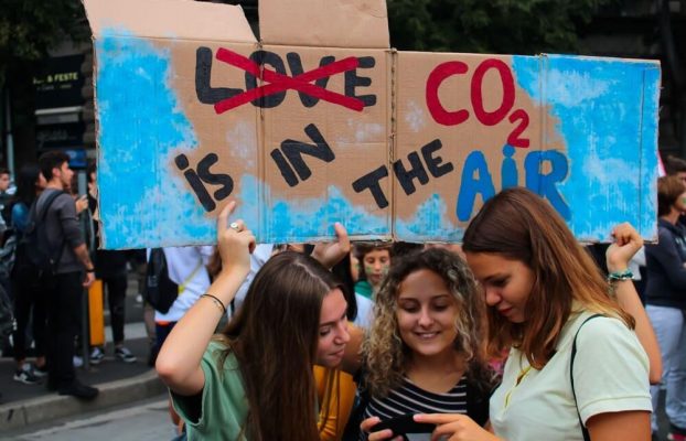 Trois jeunes filles regardent un écran de téléphone sous une pancarte que l’une d’entre elles tient. On peut lire dessus : CO2 is in the air avec le mot love barré.
