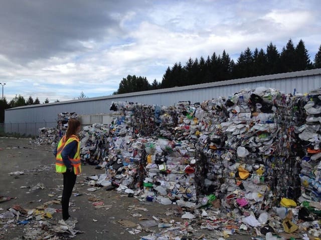 Une femme avec un gilet jaune se tient devant des monticules de déchets compressés.