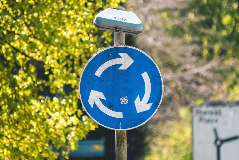 Un panneau de signalétique bleu avec des flèches en cercle sur fond arboré.