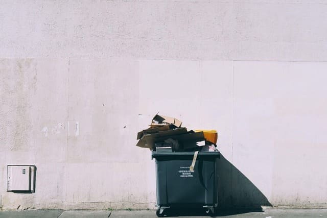 : Une poubelle noire remplie de cartons qui débordent et de déchets en tout genre est posée contre un mur blanc.